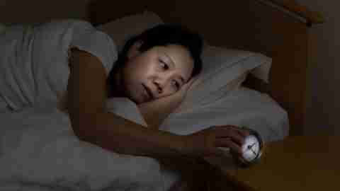 Woman with ADHD is sleepless touching alarm clock while eyes open.