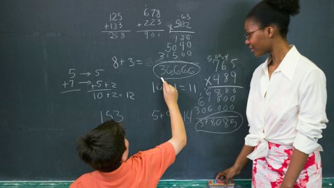 Student with ADHD writing out math problem on blackboard with teacher looking on